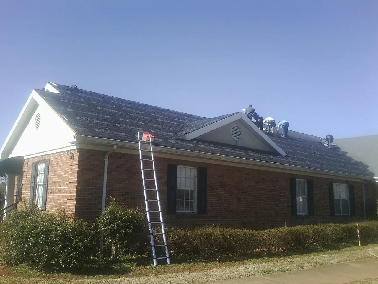 Crew tearing shingles off a brick ranch roof with a ladder set — Emergency Roof Repair work by our crew in Custar