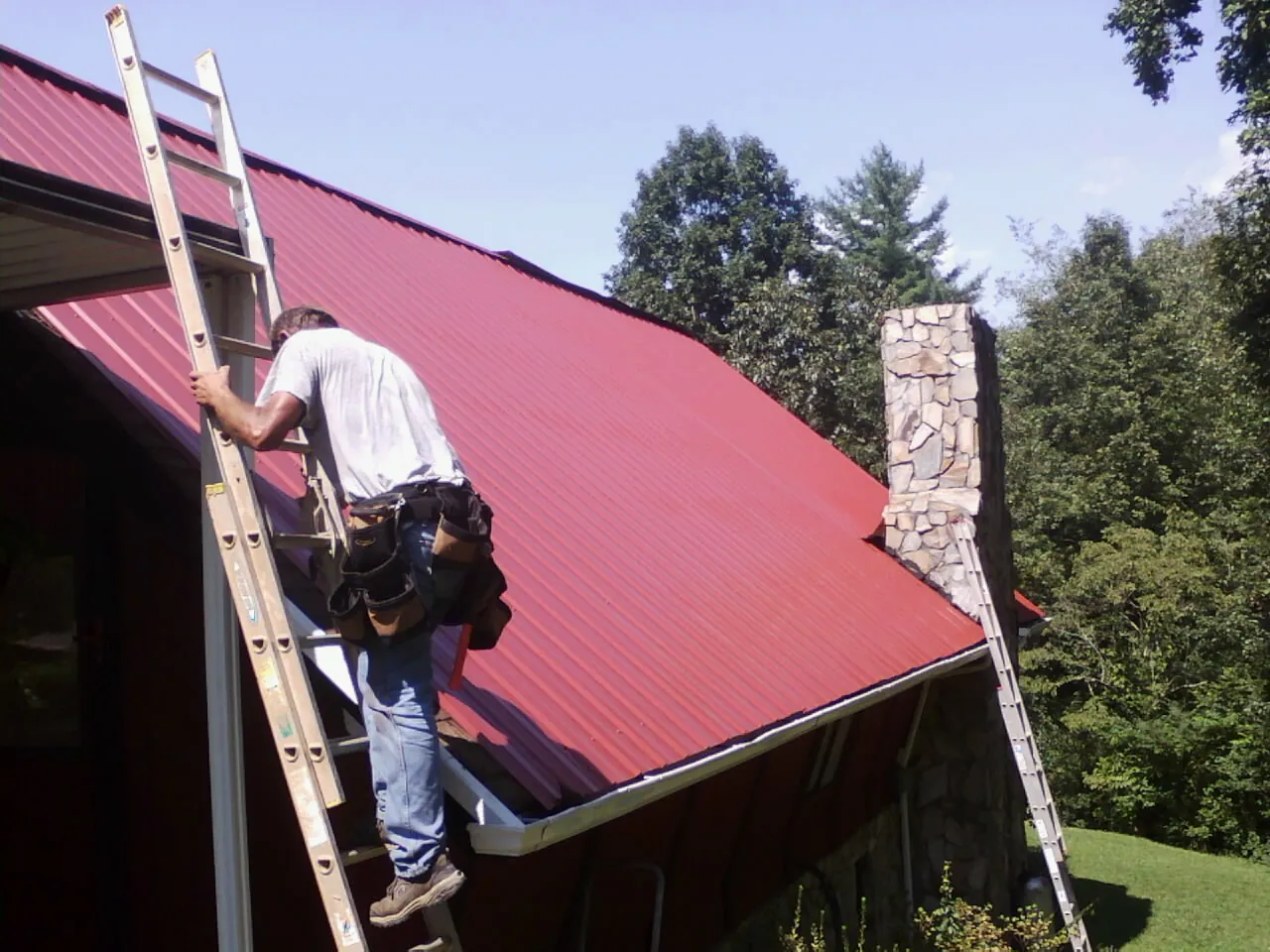 Roofer on a ladder beside a red metal roof and stone chimney — Soffit & Fascia Repair by Roofing Brothers in Custar