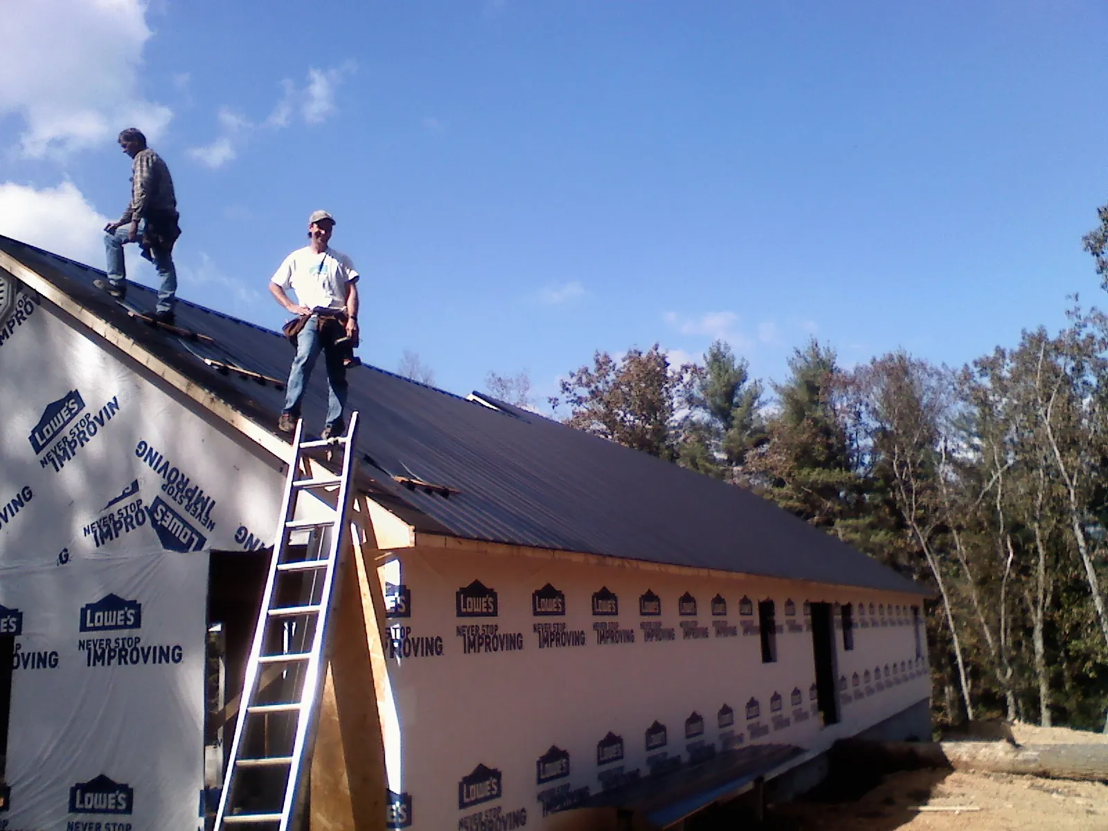 Two roofers standing on a long dark metal roof over a wrapped building in Custar